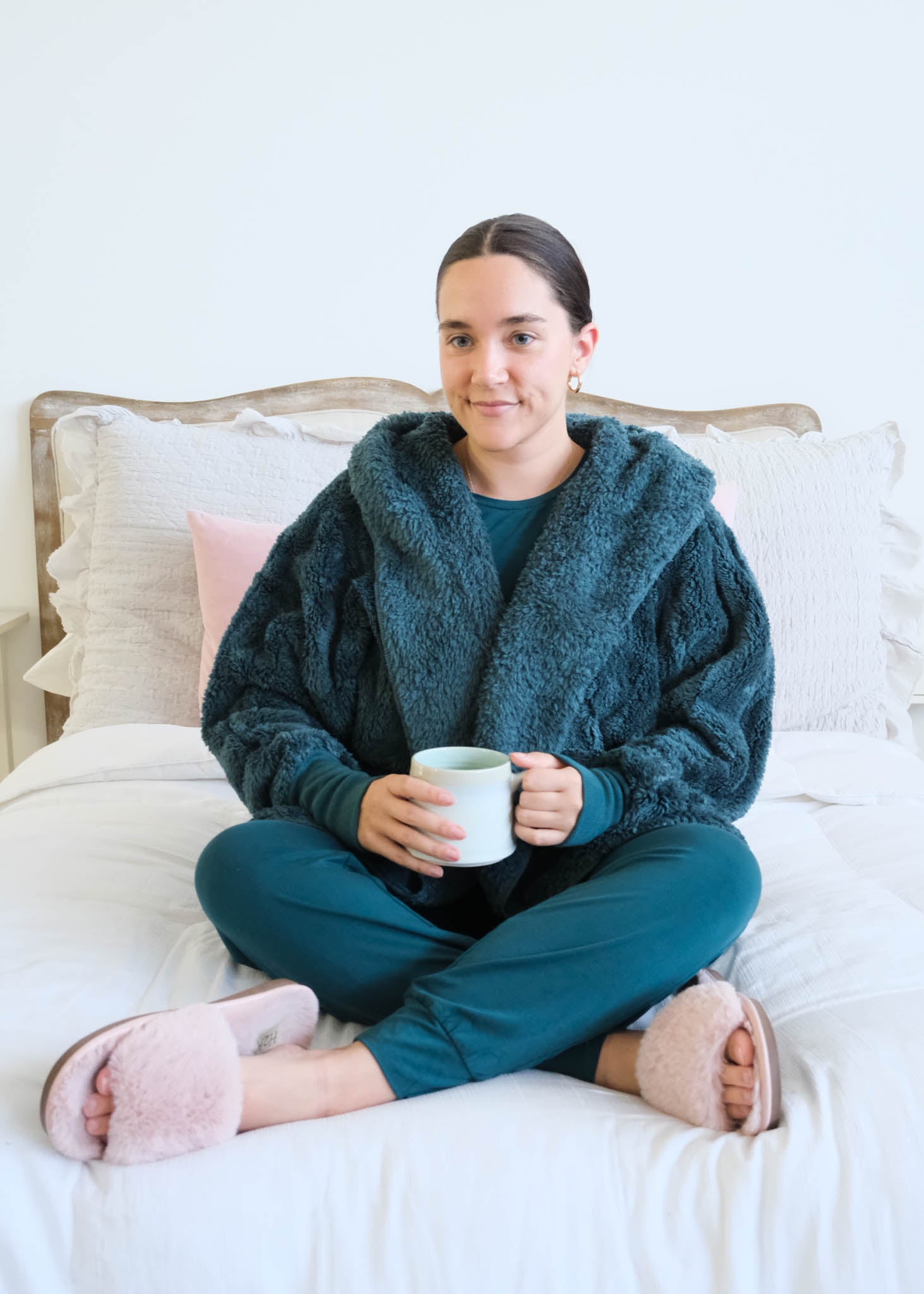 A woman sits cross-legged on a white bed wearing a cozy dark teal fuzzy hooded and matching bamboo teal pajama set, holding a white mug. She has pink slippers on her feet, with white and light pink pillows behind her. Cloud Nine Pajamas Edmonton