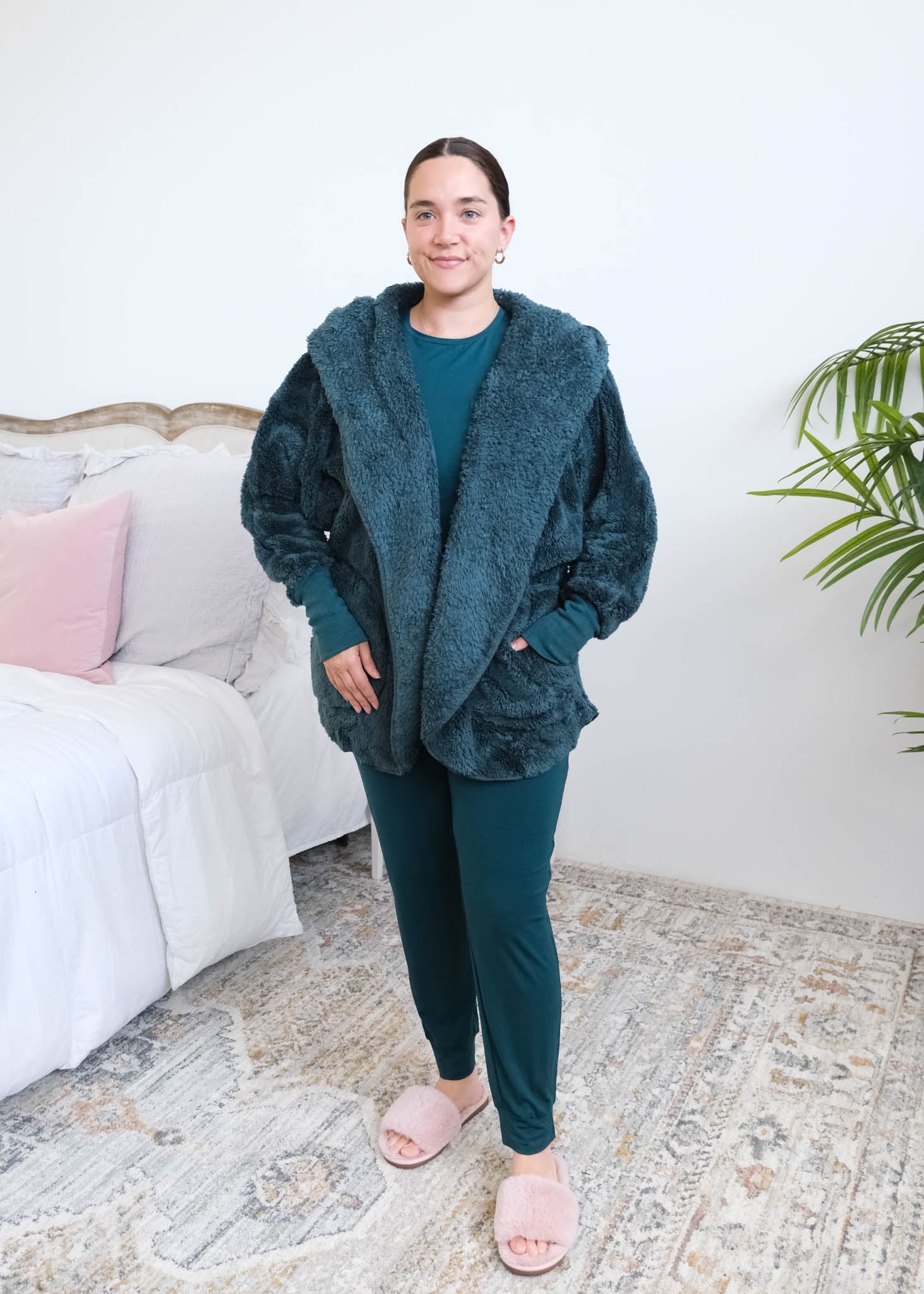 Woman standing in a bedroom wearing a dark peacock teal fluffy hooded cardigan by Lounge by Jan Marie over a dark midnight teal bamboo pajamas, with ribbed cuffs and pink fuzzy slippers, hands tucked into the cardigan's front pockets. Cloud Nine Pajamas Edmonton