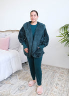 Woman standing in a bedroom wearing a dark peacock teal fluffy hooded cardigan by Lounge by Jan Marie over a dark midnight teal bamboo pajamas, with ribbed cuffs and pink fuzzy slippers, hands tucked into the cardigan's front pockets. Cloud Nine Pajamas Edmonton