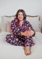 Lifestyle photo on plus size model in bedroom setting Georgia Rylan women's bamboo long sleeve button-up top and straight-leg pajama pants in Dreamy Blooms black floral print - Cloud Nine Pajamas