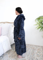 A woman wearing a plush navy blue hooded robe by Lounge By Jan Marie stands sideways in a bedroom setting with a bed, rug, and plant visible in the background. Cloud Nine Pajamas Edmonton