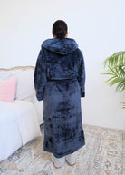 A woman wearing a plush navy blue hooded robe by Lounge By Jan Marie stands backwards in a bedroom setting with a bed, rug, and plant visible in the background. Cloud Nine Pajamas Edmonton