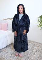 A woman wearing a plush navy blue hooded robe by Lounge By Jan Marie with the hood up on her head stands in a bedroom setting with a bed, rug, and plant visible in the background. Cloud Nine Pajamas Edmonton