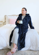 A woman sitting on a bed with a cup of coffee wearing a plush navy blue hooded robe by Lounge By Jan Marie  in a bedroom setting with a bed. Cloud Nine Pajamas Edmonton