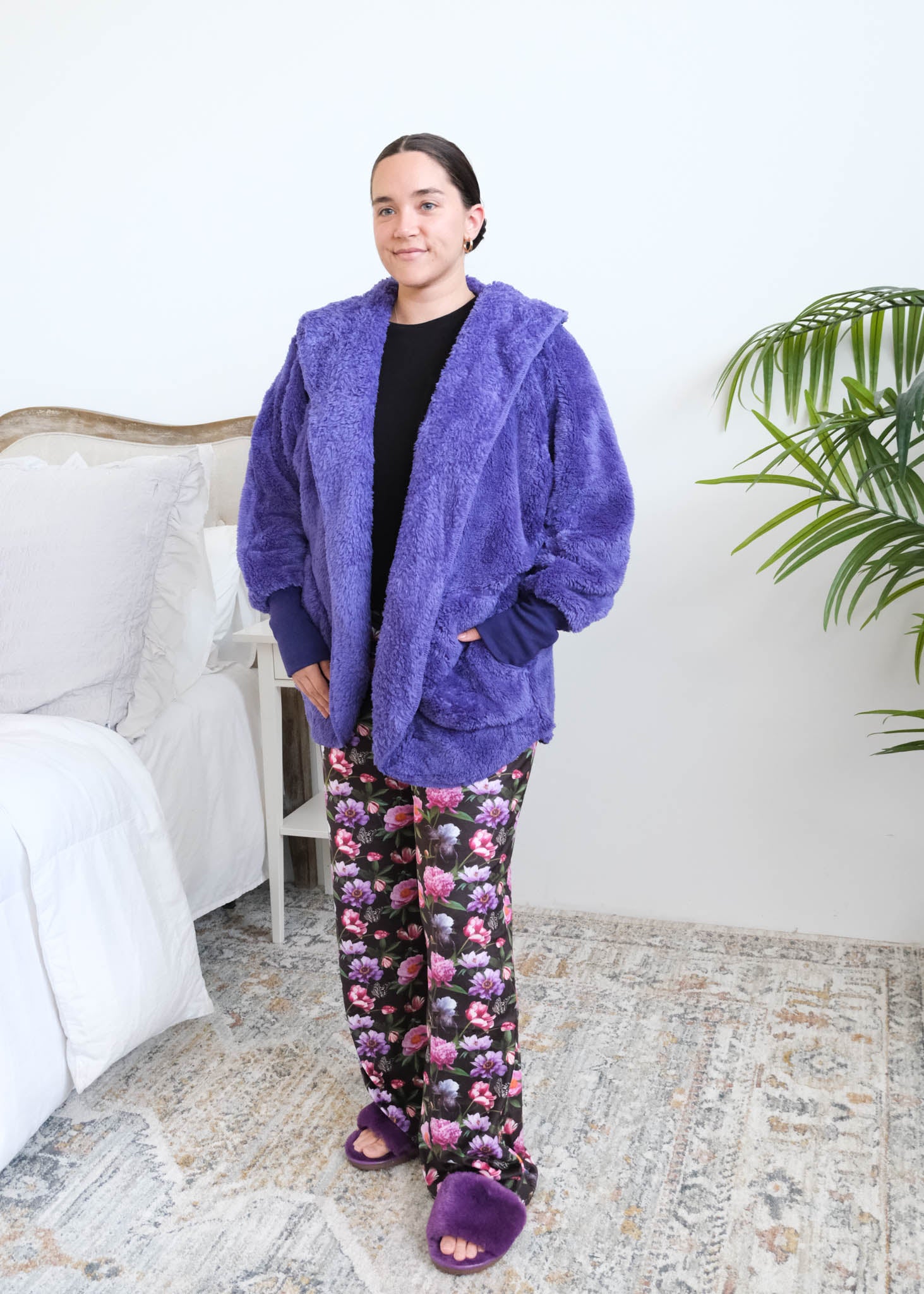 Woman standing in a bedroom wearing a blueberry purple fluffy hooded cardigan over a black top and floral pajama pants, with purple ribbed cuffs and purple fuzzy slippers, hands tucked into the cardigan's front pockets. Cloud Nine Pajamas Edmonton