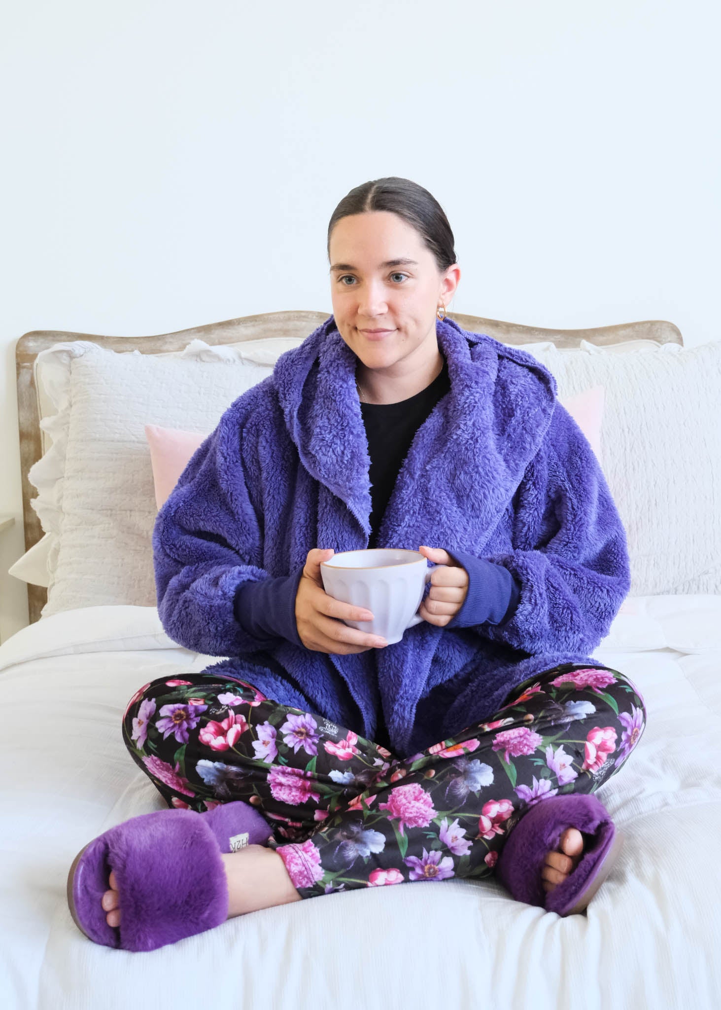 A woman sits cross-legged on a white bed wearing a flully  plush sherpa purple hooded  cardigan by Lounge by Jan Marie, floral pajama pants, and matching purple slippers while holding a mug. Cloud Nine Pajamas Edmonton 