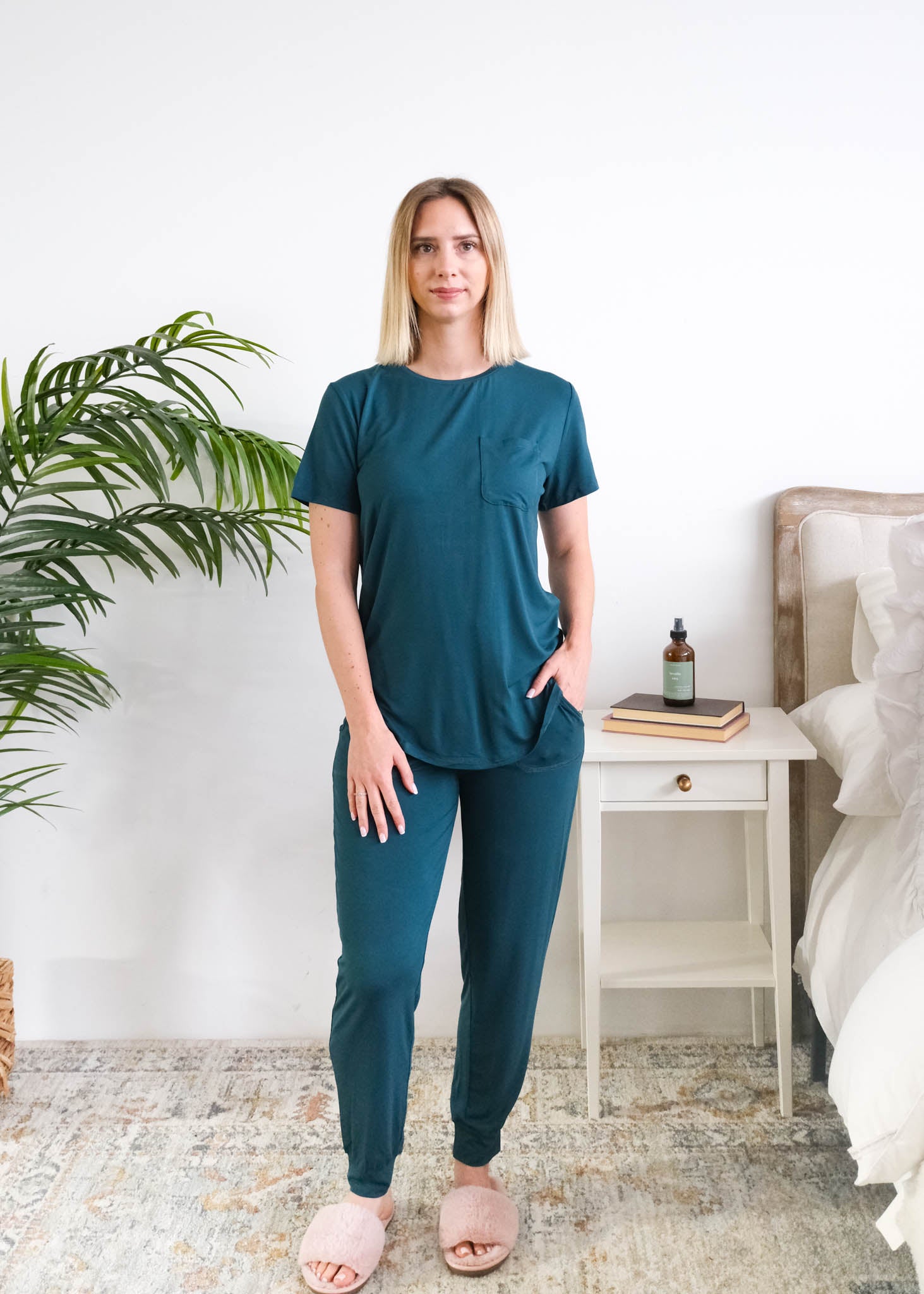 A woman modelling a matching two-piece bamboo dark teal loungewear set in a bright bedroom setting. The set consists of a short-sleeve crewneck top and matching jogger pants in a deep teal color. Cloud Nine Pajamas Edmonton