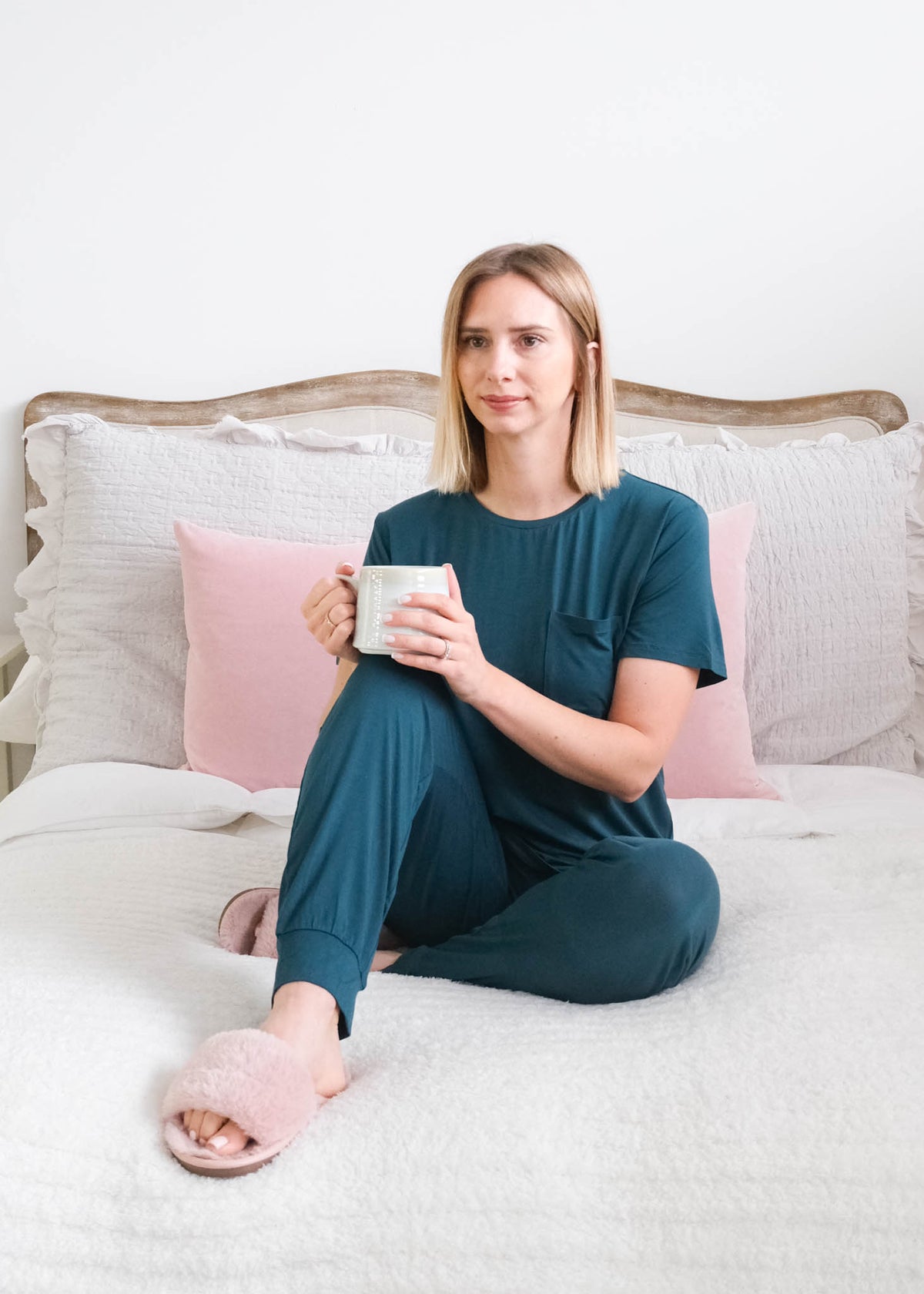 A woman sits on a bed wearing a dark teal short-sleeve pajama set with jogger style bottoms. She is wearing pink slippers, holding a white mug. The background features a white headboard, white bedding, and pink pillows. Cloud Nine Pajamas Edmonton