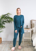 A woman modeling a matching bamboo dark teal lounge set consisting of a long-sleeve open-front cardigan, a long-sleeve top, and jogger pants, styled in a bright bedroom setting. Cloud Nine Pajamas Edmonton