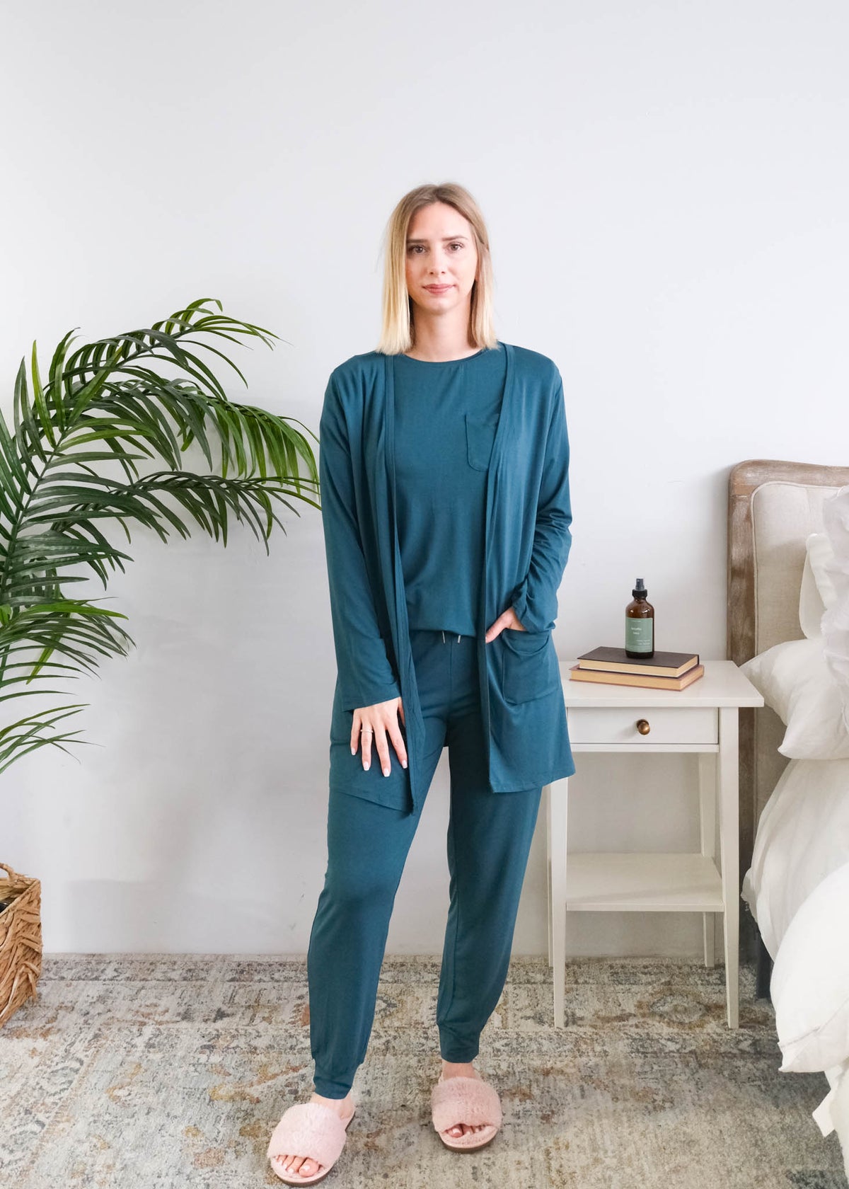 A woman modeling a matching bamboo dark teal lounge set consisting of a long-sleeve open-front cardigan, a long-sleeve top, and jogger pants, styled in a bright bedroom setting. Cloud Nine Pajamas Edmonton