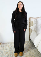 Model with long dark hair wearing a black long-sleeve scoop-neck pajama top half-tucked into matching black wide-leg pants with a drawstring tie waist, standing in a bedroom beside a white bed, styled with tan platform slippers. Cloud Nine Pajamas Edmonton. 