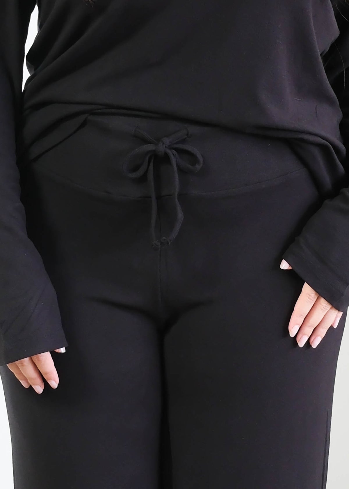 Close-up of black wide-leg pajama pants showing the wide, smooth fold-over waistband with a functional drawstring tied in a bow, worn with the matching black long-sleeve top. Cloud Nine Pajamas Edmonton. 