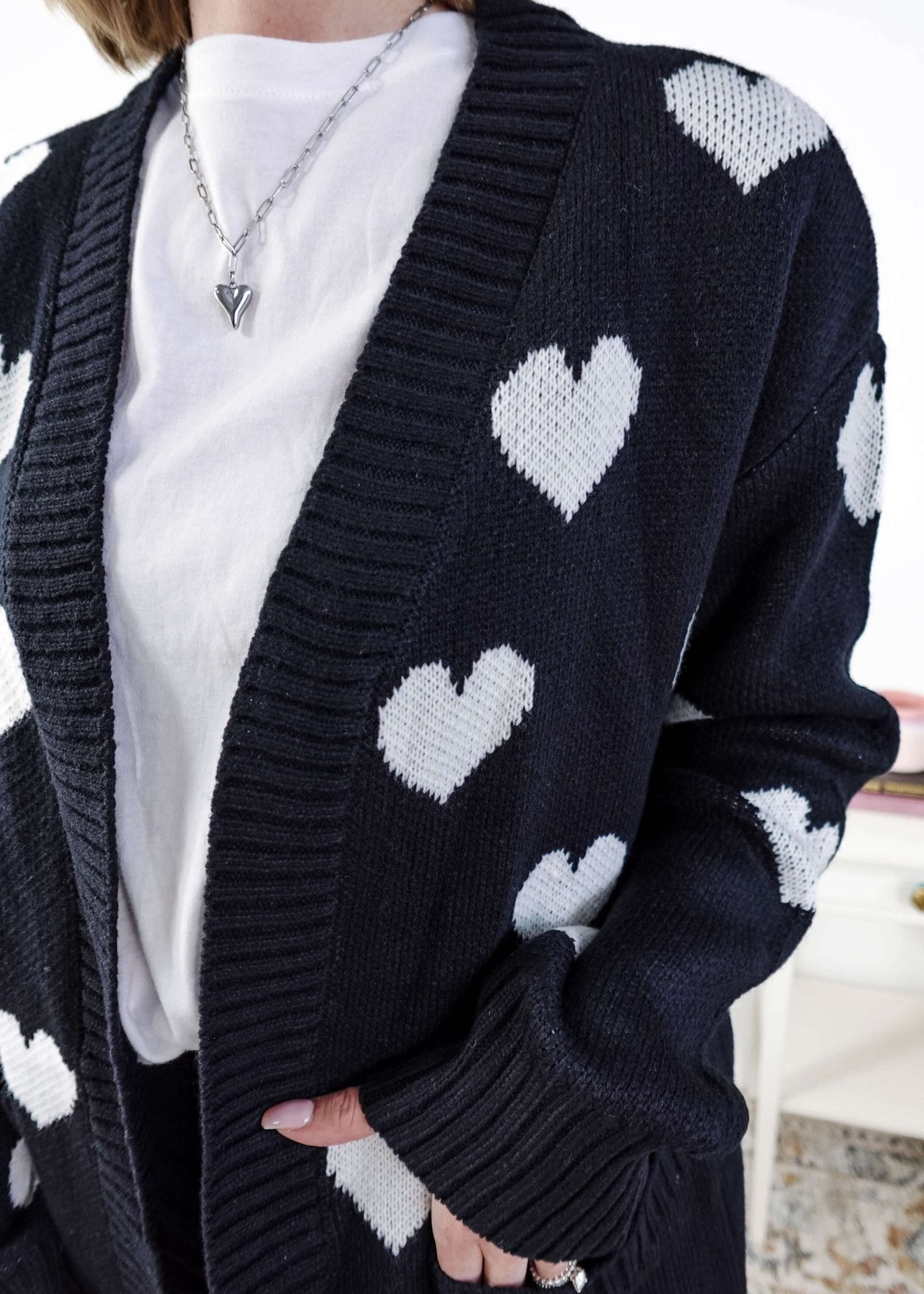 Scarlett Ellie Black Hearts Knit Cardigan | Cloud Nine Pajamas Women standing in a long black open cardigan with white hearts on it in a bedroom 