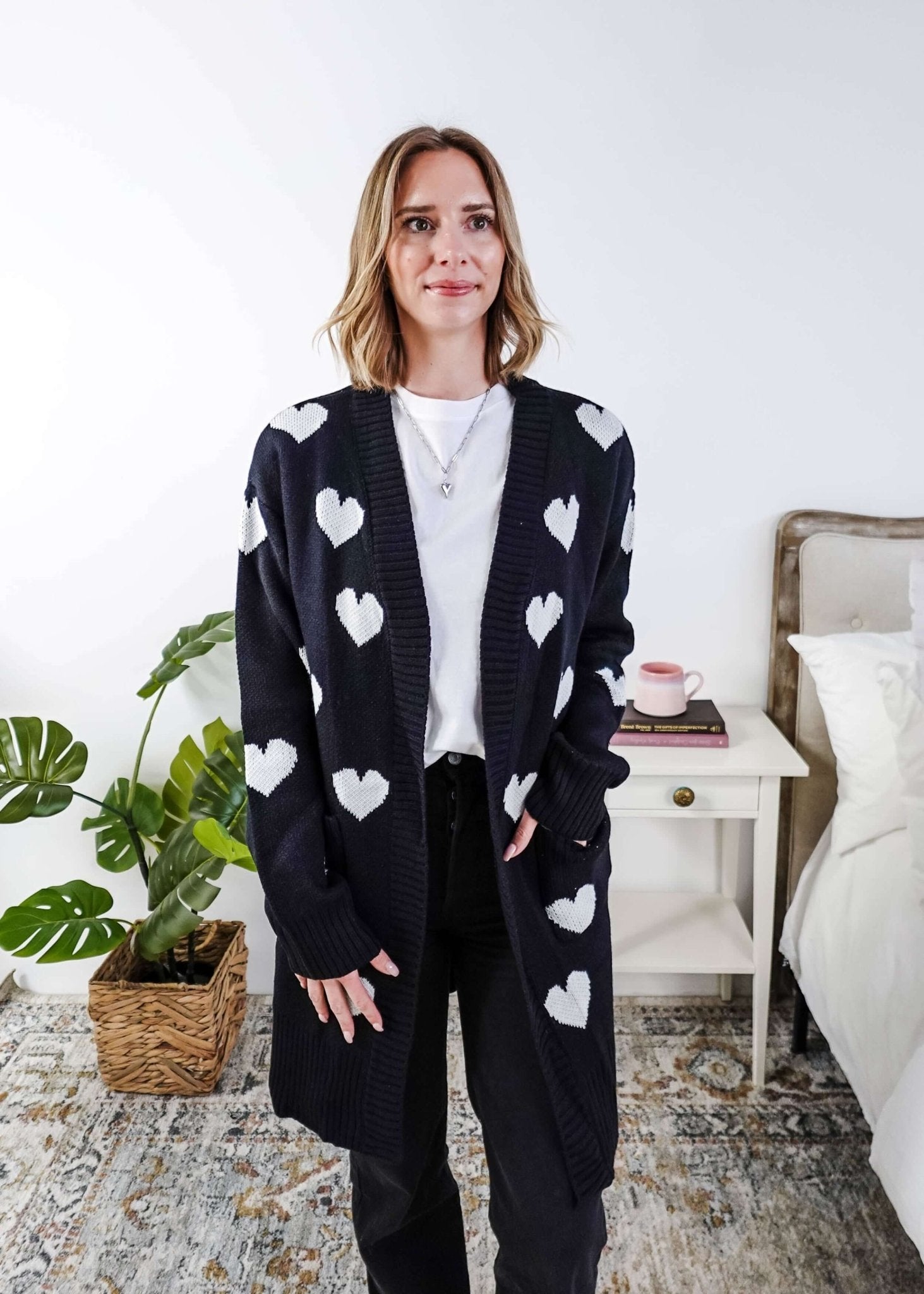 Scarlett Ellie Black Hearts Knit Cardigan | Cloud Nine Pajamas - Women standing in a long black open cardigan with white hearts on it in a bedroom 