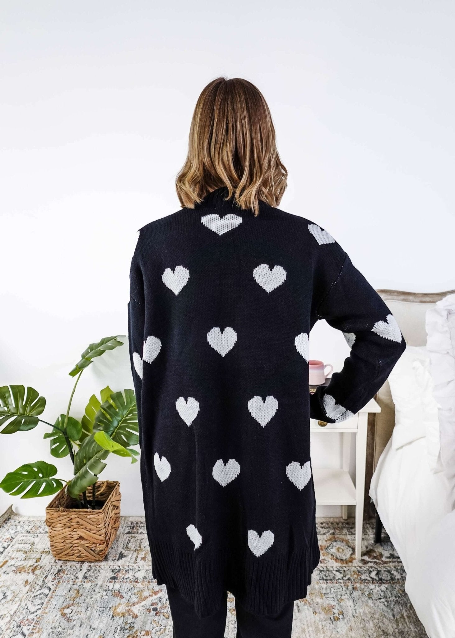 Women standing in a long black open cardigan with white hearts on it in a bedroom showing back of cardigan and the striking white hearts on a black background - Scarlett Ellie Black Hearts Knit Cardigan | Cloud Nine Pajamas