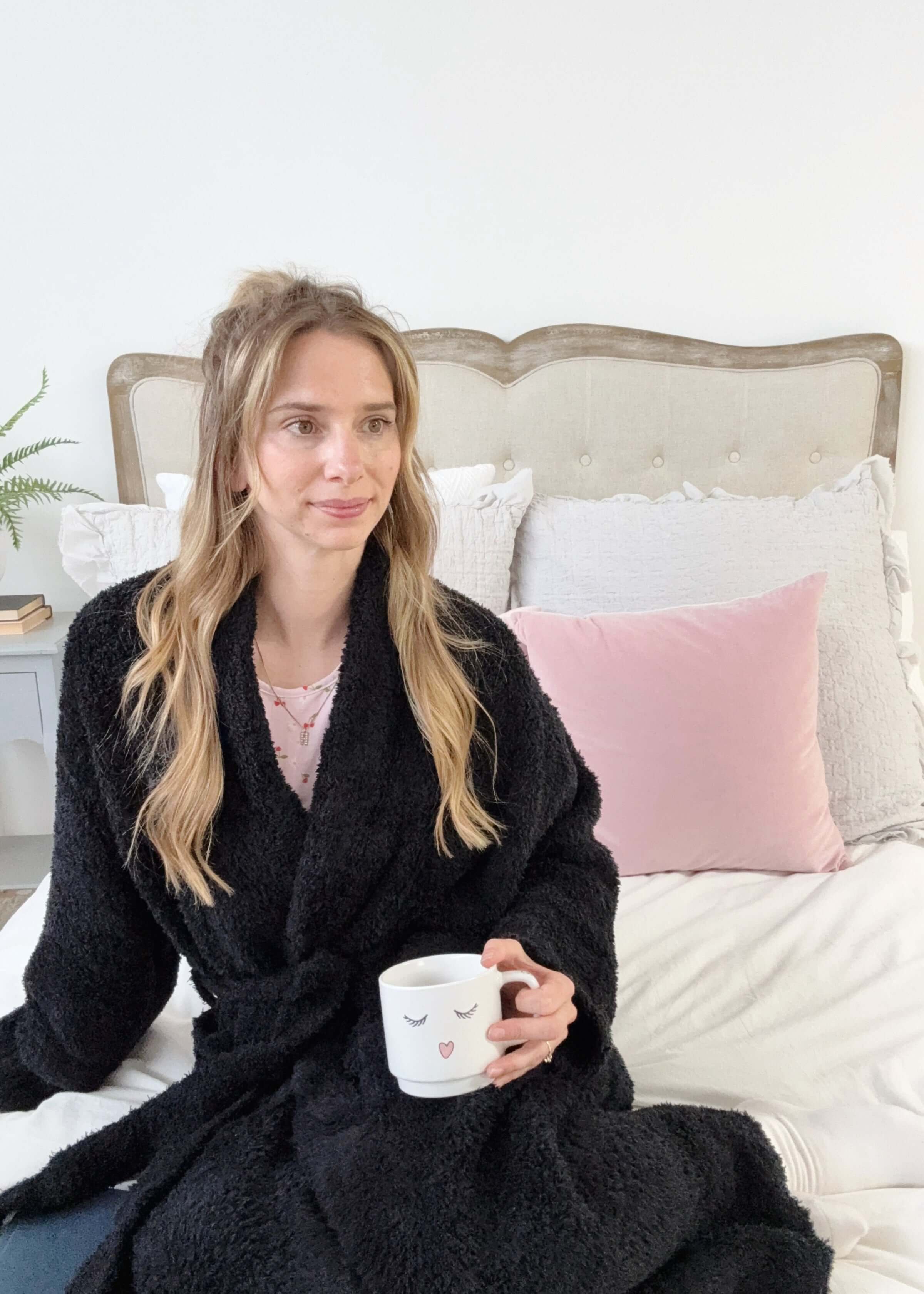 Woman in a black robe holding a mug on a bed with pink and gray pillows.