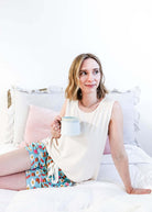 Lifestyle shot of Miranda on a bed wearing the Brunch Club velour shorts with eggs and bacon print and ivory peachy tank.
