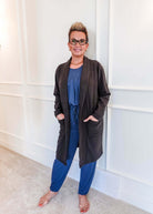 Full-length photo of a woman smiling, standing in a hallway, wearing a Charcoal Elevate Cardigan with deep patch pockets layered over a blue pyjama set. Classic collar, long-line structured fit.