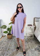 Woman in a light purple dress standing in a bedroom.