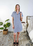 Woman in a blue and white striped dress holding a brown bag in a bedroom setting. Scarlett Ellie Collared Button-Up Short Sleeve Dress | Cloud Nine Pajamas