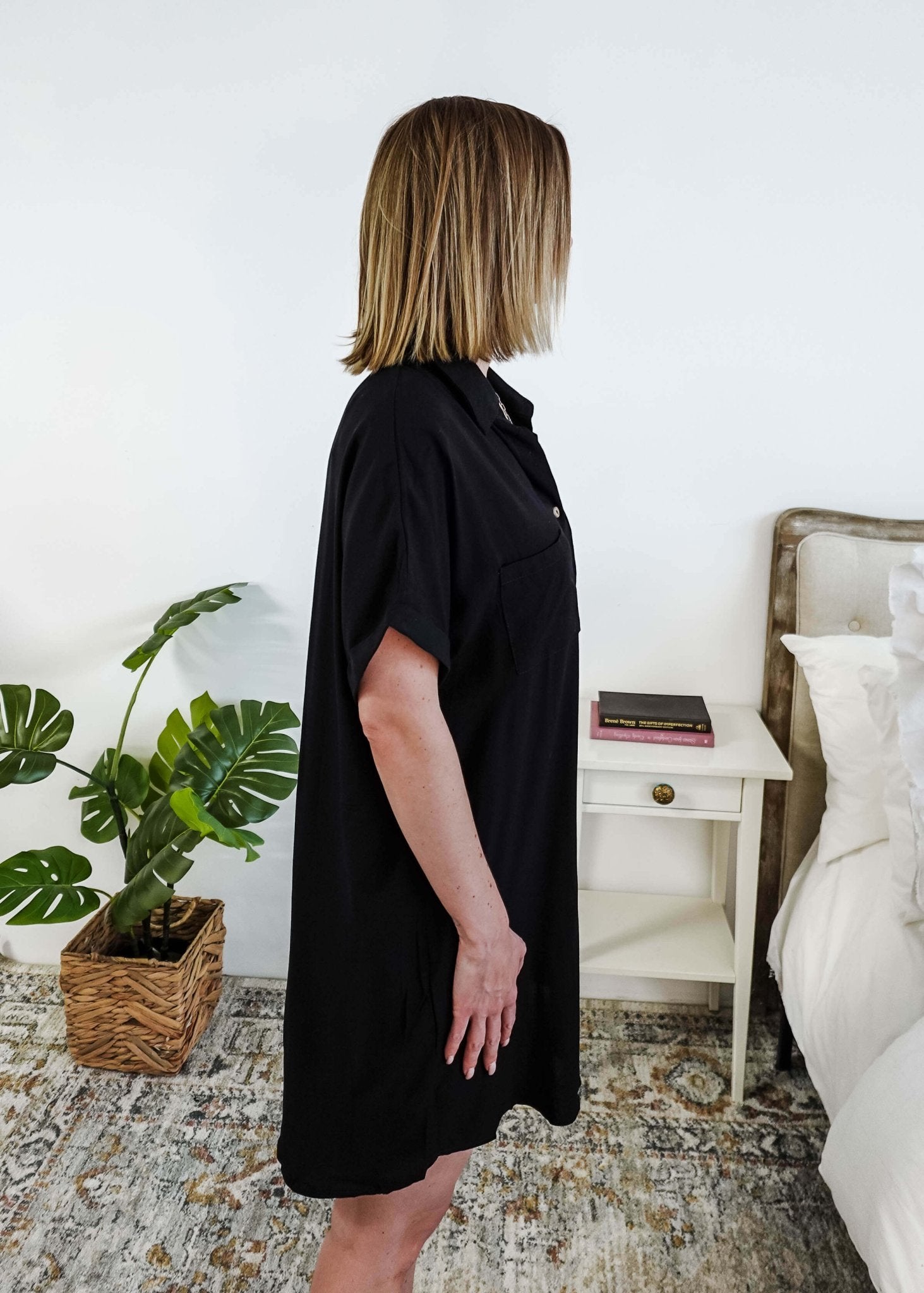 Woman wearing a black dress standing in a bedroom with a plant and nightstand. Scarlett Ellie Collared Button-Up Short Sleeve Dress | Cloud Nine Pajamas