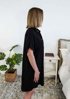 Woman wearing a black dress standing in a bedroom with a plant and nightstand. Scarlett Ellie Collared Button-Up Short Sleeve Dress | Cloud Nine Pajamas