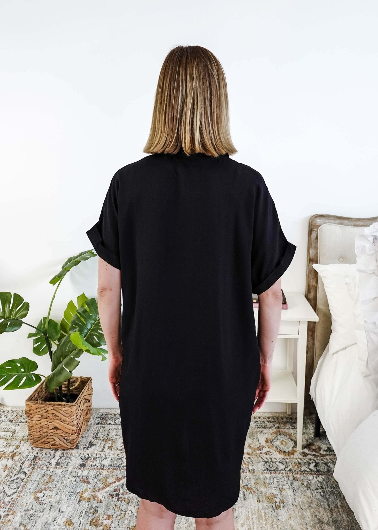 Person wearing a black dress standing in a bedroom with a plant and bed in the background Scarlett Ellie Collared Button-Up Short Sleeve Dress | Cloud Nine Pajamas showing back view