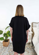 Person wearing a black dress standing in a bedroom with a plant and bed in the background Scarlett Ellie Collared Button-Up Short Sleeve Dress | Cloud Nine Pajamas showing back view