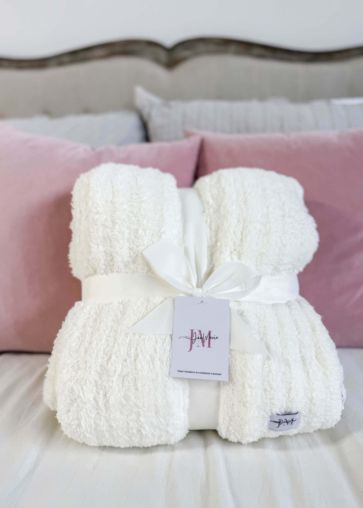 White fluffy blanket with a tag on a bed with pink pillows. Cozy Cabin Blanket – Oversized Ivory Plush Throw (75″ × 52″)