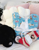 Dog Lover Gift Bundle Dog-themed gift set with a blue pj set with dogs skating print, red and beige pug socks, and a black plush labrador warming heat pack and hand rescue cream on a soft surface.
