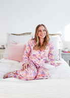 Woman sitting on bed in Dream in Blooms Long Sleeve Button Up & Pant Bamboo Lounge Set with floral design.