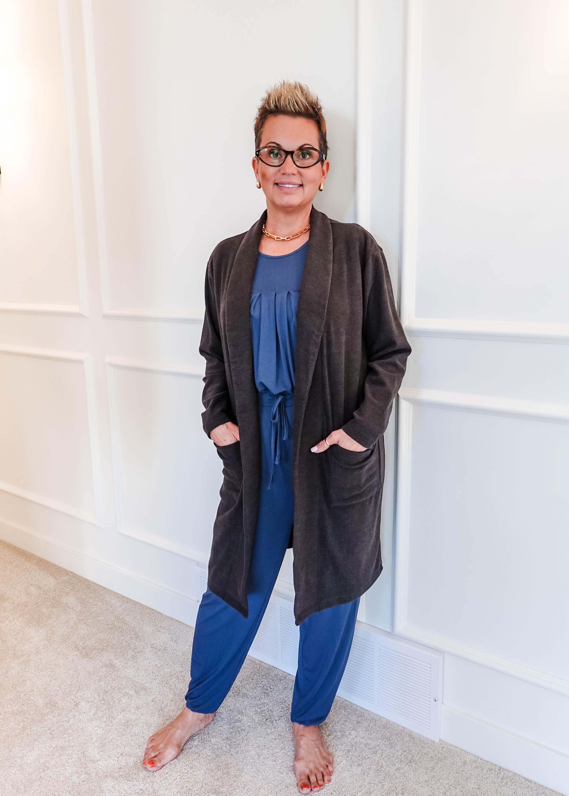Full-length photo of a woman smiling, standing in a hallway, wearing a Charcoal Elevate Cardigan with deep patch pockets layered over a blue pyjama set. Classic collar, long-line structured fit.