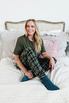 Woman sitting on a bed wearing Green Plaid Unisex Bamboo Jogger from Bob's Sleepwear Co. in a cozy bedroom setup.