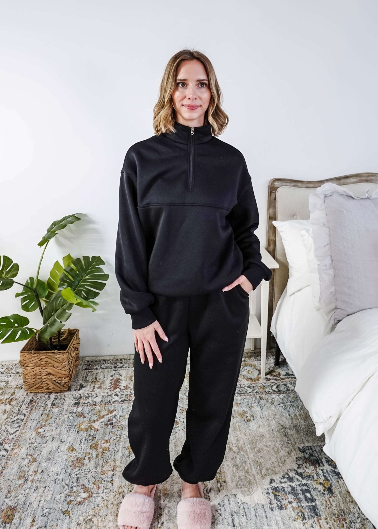 Woman wearing a black sweatsuit standing in a bedroom with a plant and bed in the background, Scarlett Ellie Half-Zip Sweatsuit Set | Cloud Nine Pajamas