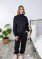 Woman wearing a black sweatsuit standing in a bedroom with a plant and bed in the background, Scarlett Ellie Half-Zip Sweatsuit Set | Cloud Nine Pajamas