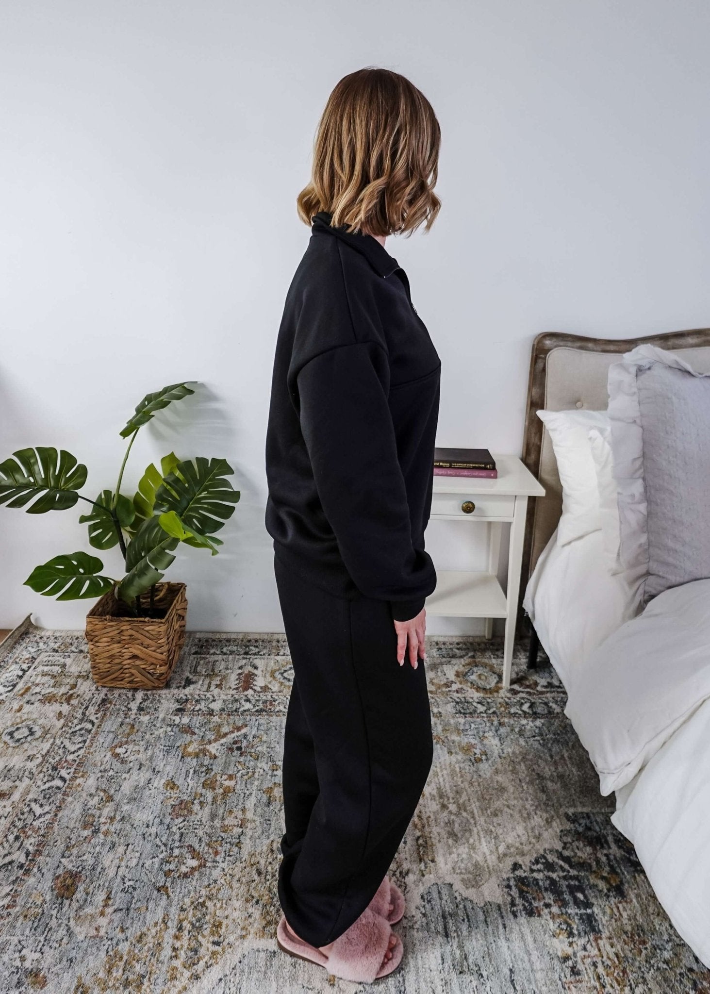 Woman wearing a black sweatsuit standing in a bedroom with a plant and bed in the background, Scarlett Ellie Half-Zip Sweatsuit Set | Cloud Nine Pajamas