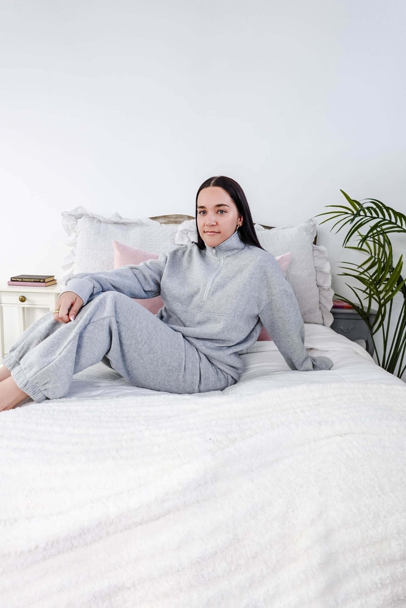 Person wearing a gray outfit sitting on a bed in a room with a plant. Scarlett Ellie Half-Zip Sweatsuit Set | Cloud Nine Pajamas