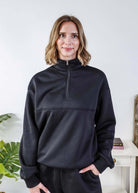 Woman wearing a black sweatsuit standing in a bedroom with a plant and bed in the background, Scarlett Ellie Half-Zip Sweatsuit Set | Cloud Nine Pajamas