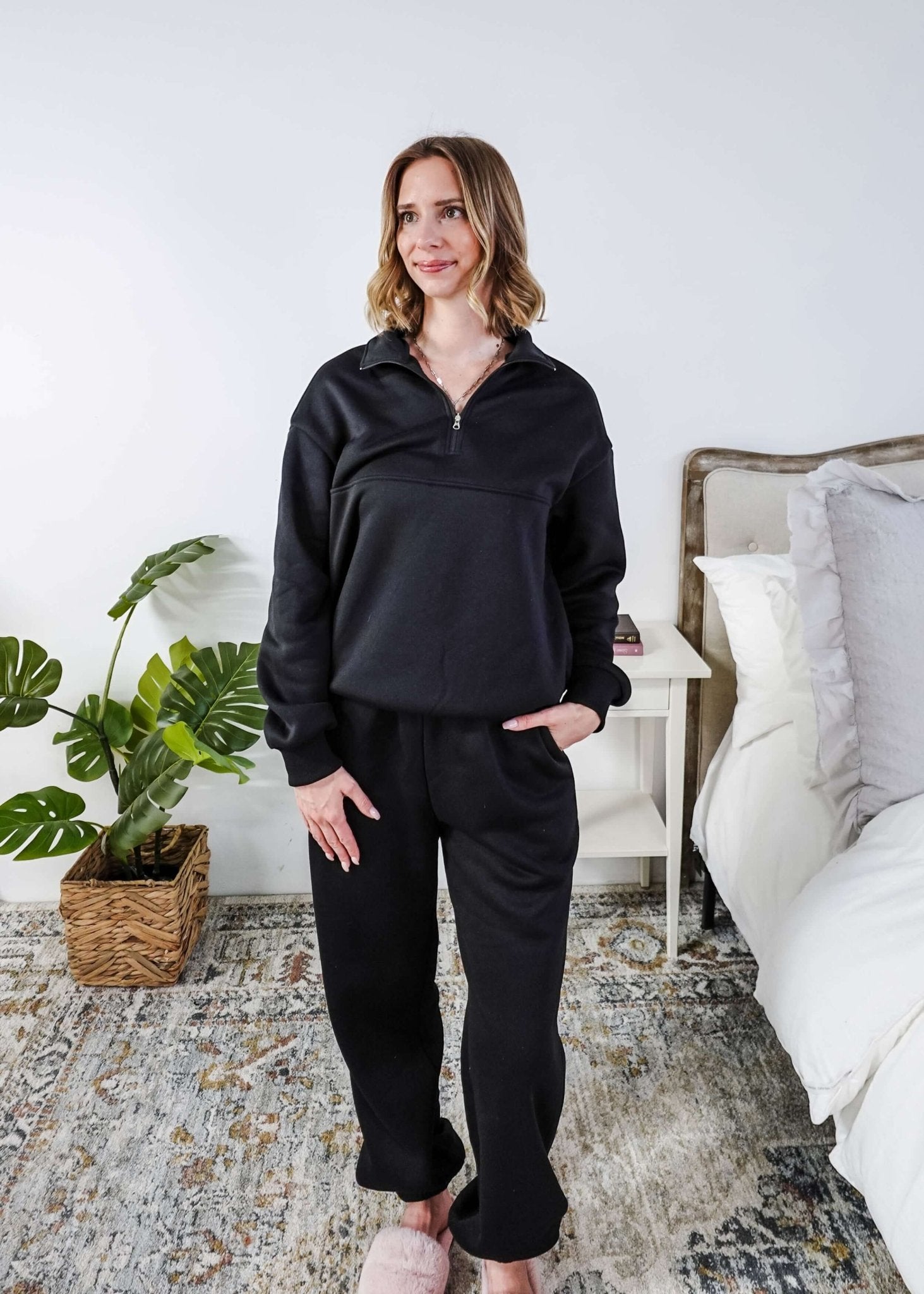 Woman wearing a black sweatsuit standing in a bedroom with a plant and bed in the background, Scarlett Ellie Half-Zip Sweatsuit Set | Cloud Nine Pajamas