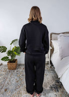 Woman wearing a black sweatsuit standing in a bedroom with a plant and bed in the background, Scarlett Ellie Half-Zip Sweatsuit Set | Cloud Nine Pajamas