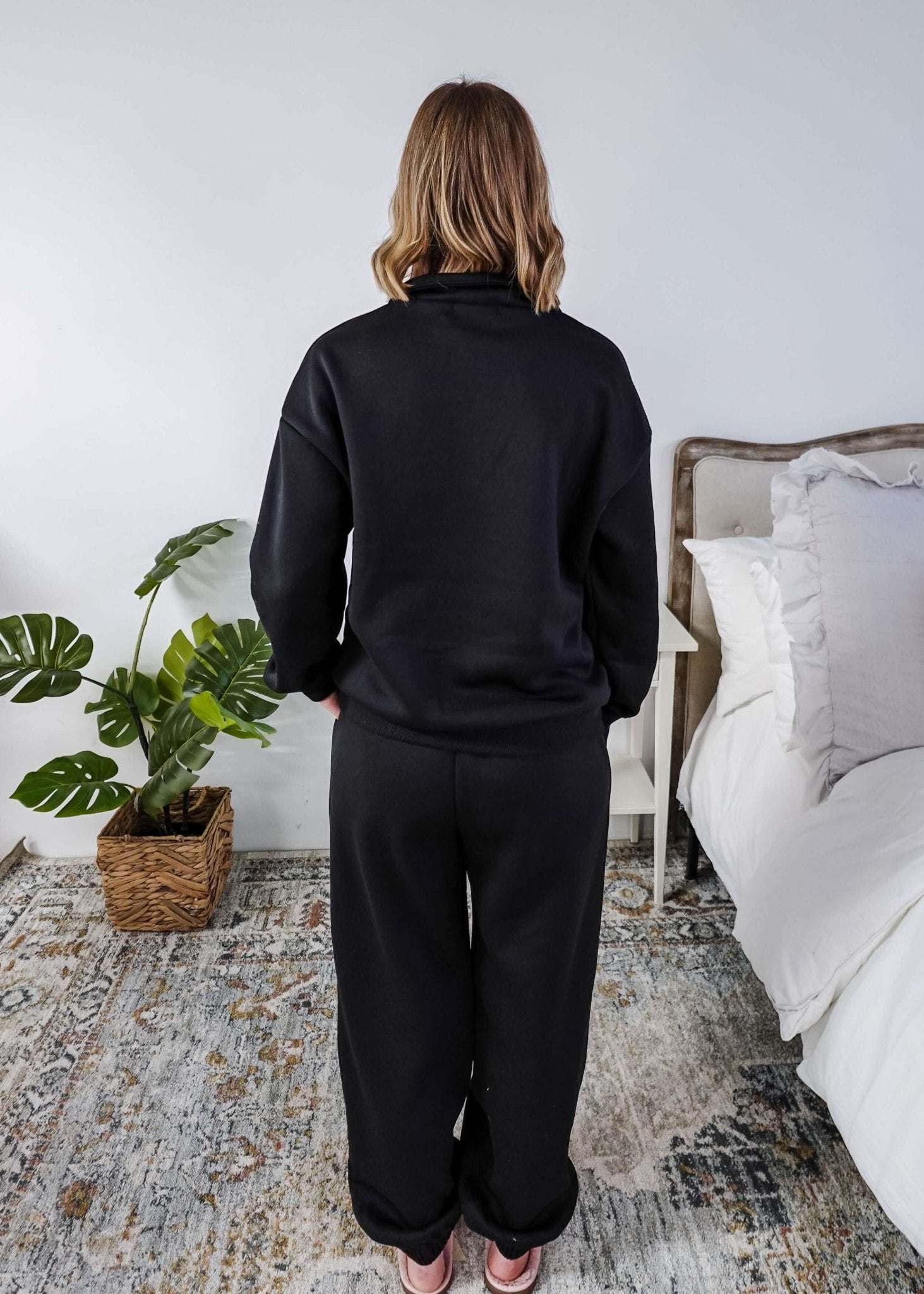 Woman wearing a black sweatsuit standing in a bedroom with a plant and bed in the background, Scarlett Ellie Half-Zip Sweatsuit Set | Cloud Nine Pajamas