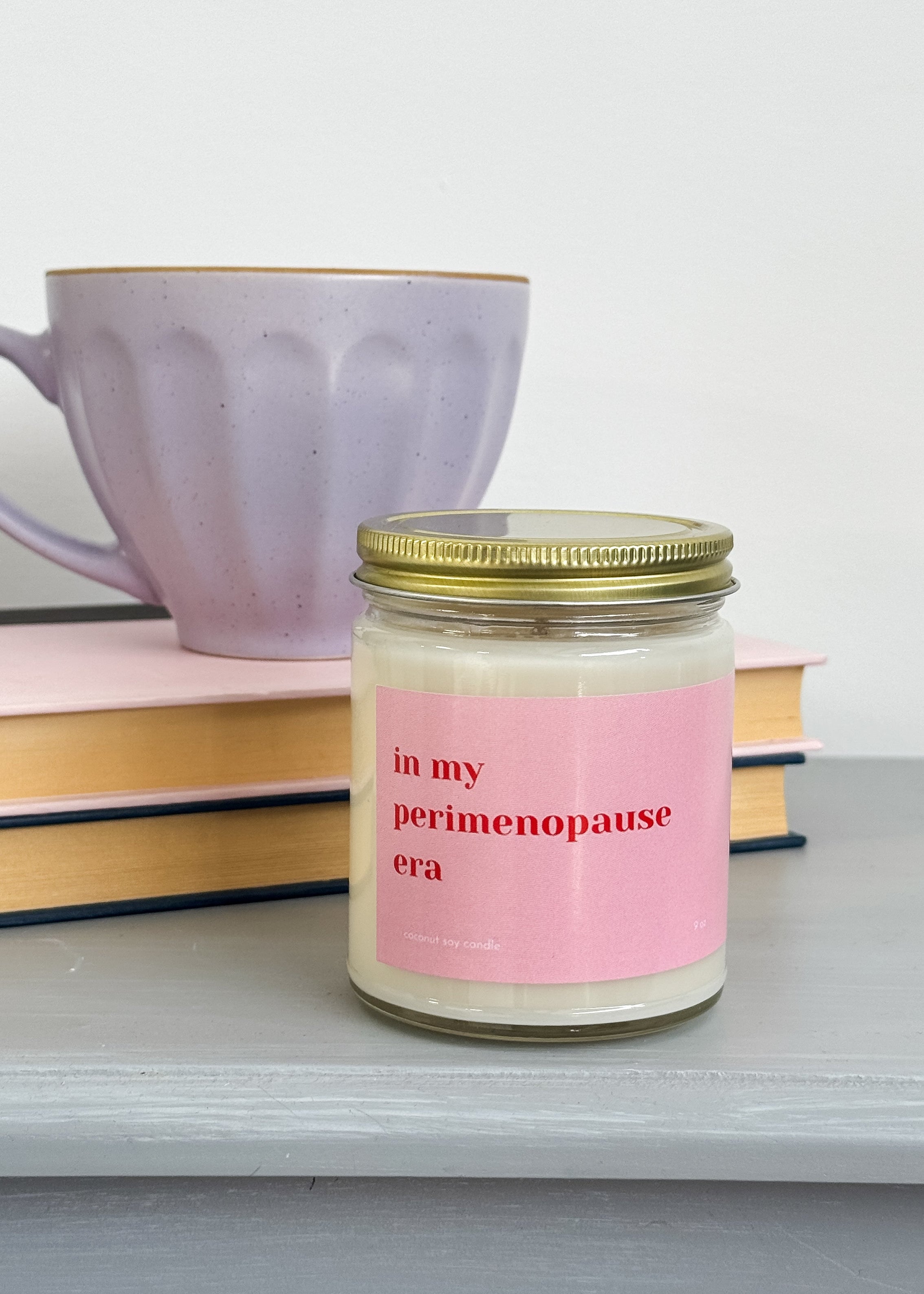 Jar of candle with 'in my perimenopause era' label on a surface with books and a mug by Heslip Home