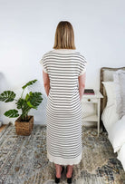 Woman wearing a black andwhite striped dress standing in a bedroom with a plant and bed in the background.