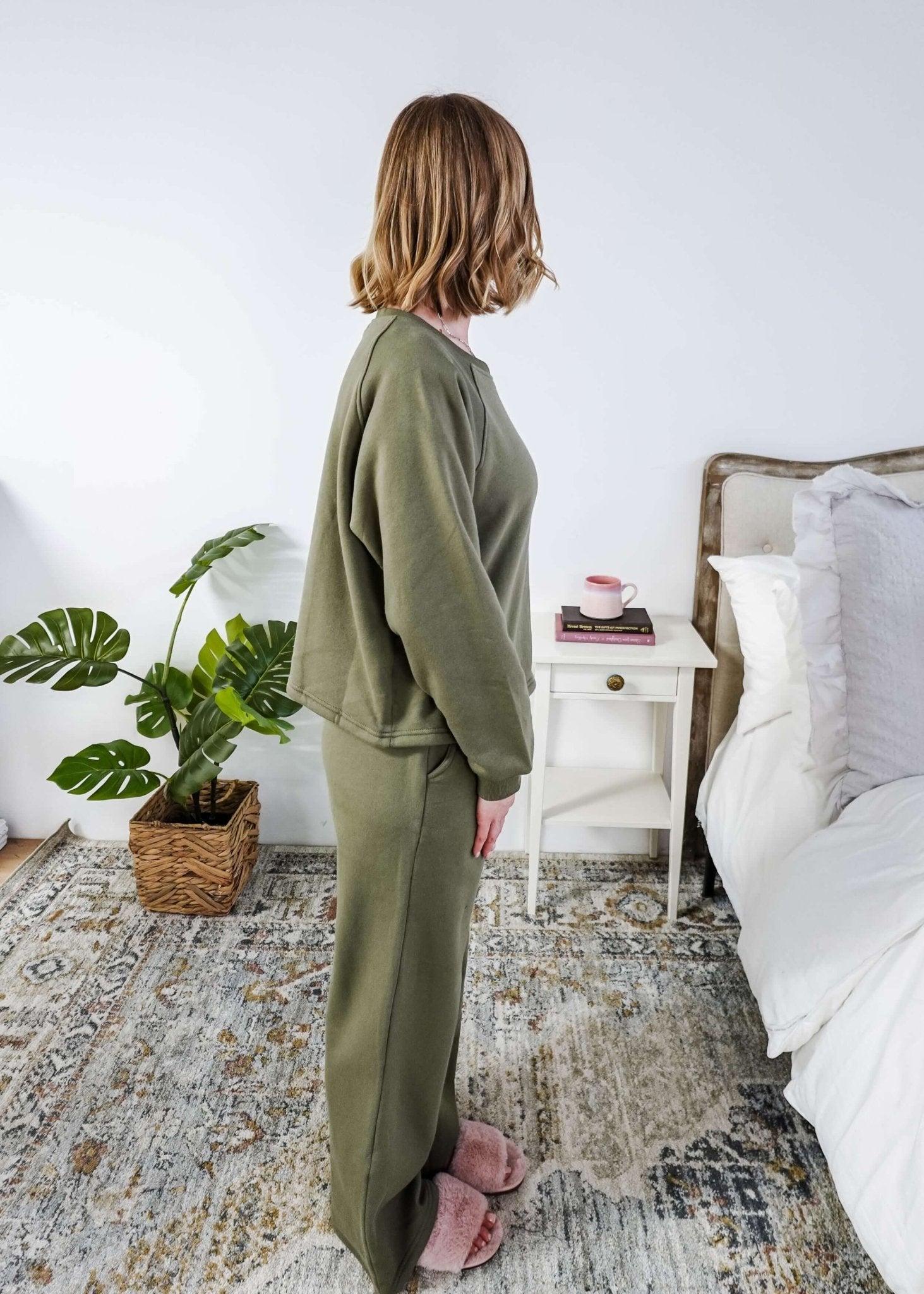 Woman in green pajama set standing in a bedroom with a plant and nightstand.