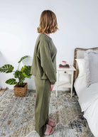 Woman in green pajama set standing in a bedroom with a plant and nightstand.