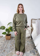 Woman wearing a green sweatsuit outfit standing in a bedroom with a plant and bed in the background.