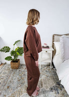 Person wearing a brown lounge outfit standing in a bedroom with a plant and bed in the background.