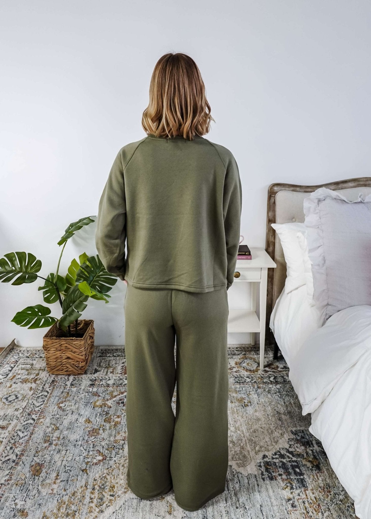 Person wearing a green outfit standing in a bedroom with a bed and plant.