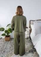 Person wearing a green outfit standing in a bedroom with a bed and plant.