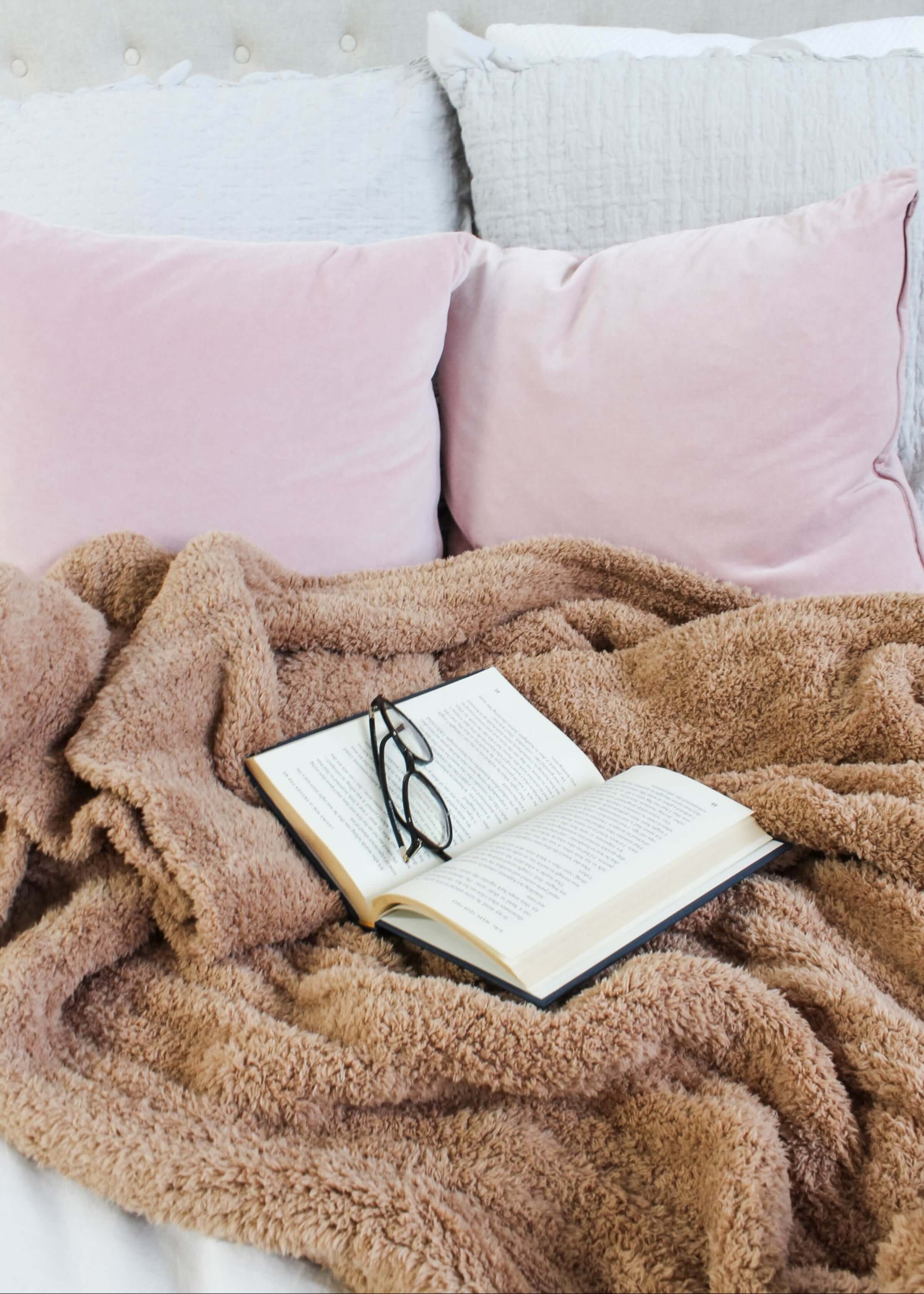 Open book with glasses on a fluffy brown blanket on a bed with pink pillows.